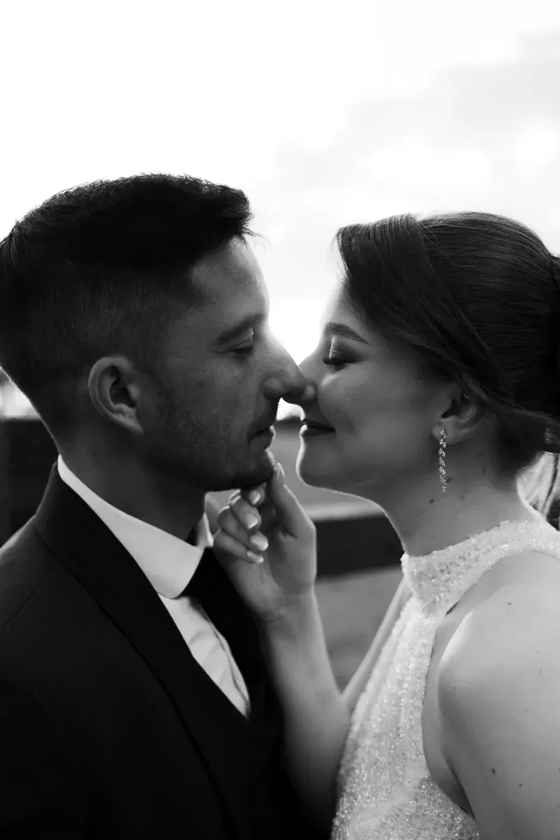 Crno-bijela intimna fotografija mladenaca nos uz nos u zalasku sunca kraj drvene ograde.
