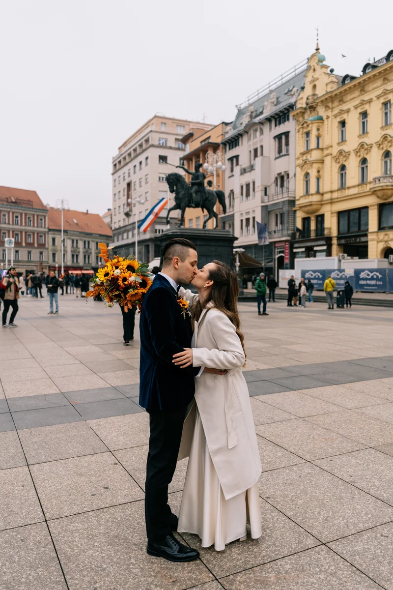Romantičan poljubac mladenaca na Trgu bana Jelačića u Zagrebu, s prepoznatljivim kipom bana i konja u pozadini.