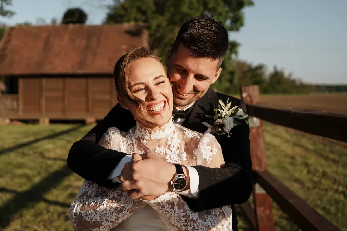 Mladoženja zagrljajem iznenađuje nasmijanu mladenku kraj drvene ograde u zlatnom svjetlu.
